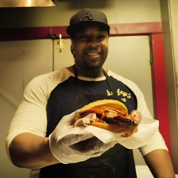 A smiling chef in an Ashley's Bar-B-Que apron holds out a delicious pulled pork sandwich.