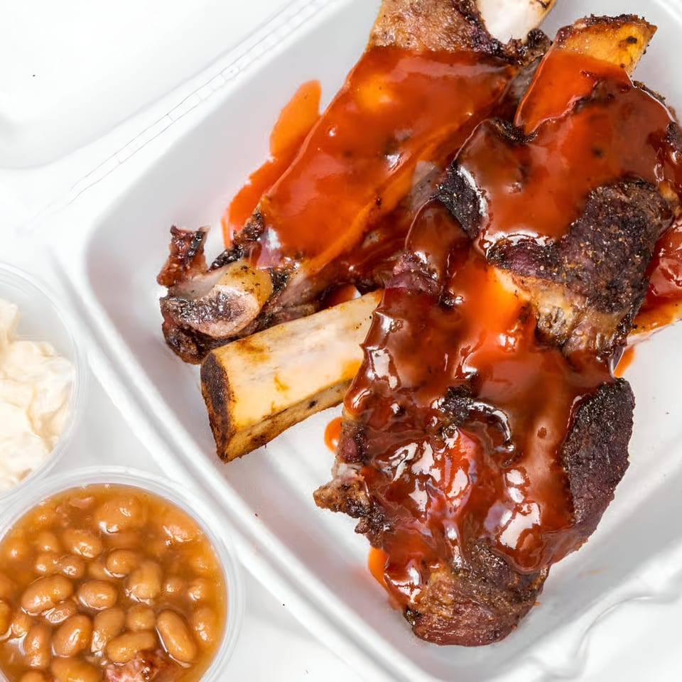 Ashley's Bar-B-Que ribs with sauce, baked beans, and potato salad in a takeout container.