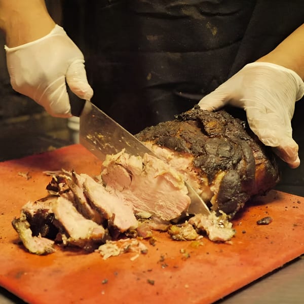 Gloved hands slicing a large, dark-crusted piece of Ashley's Bar-B-Que meat on a red cutting board.