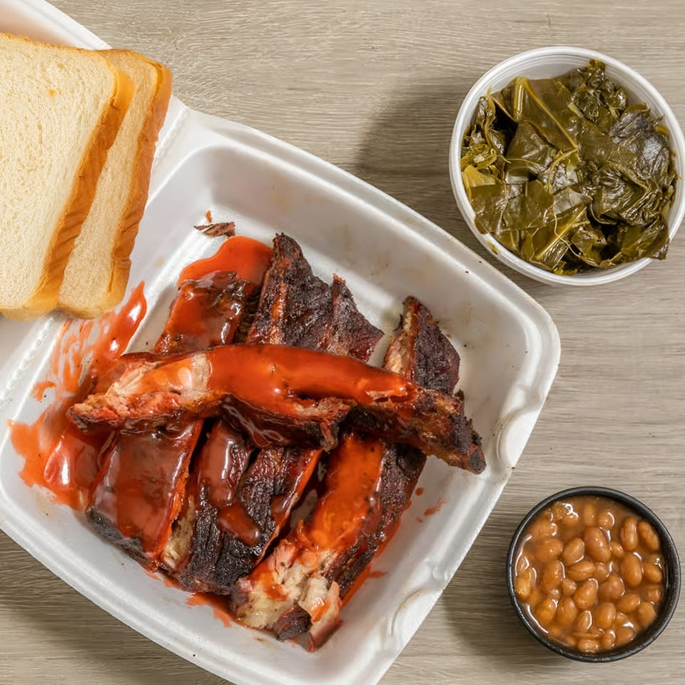 Ashley's Bar-B-Que ribs with sauce, collard greens, baked beans, and white bread in takeout containers.