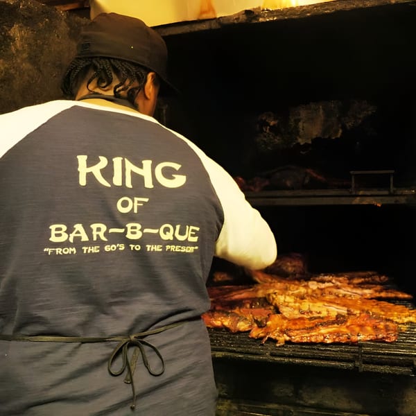 A person wearing a "King of Bar-B-Que" shirt tending to ribs on a large grill.